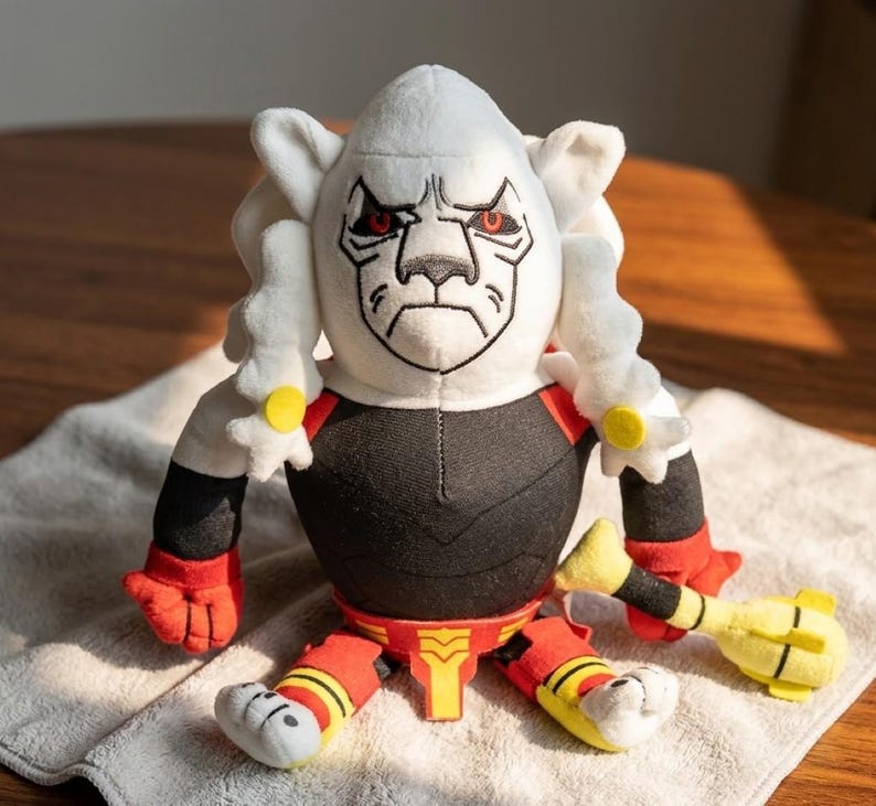 May include: A plush toy featuring a white head with a stern expression and red eyes. The toy has a black torso, red and yellow accents, and a yellow club. The toy is sitting on a white towel.
