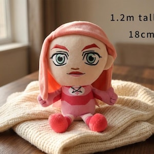 May include: A plush doll with a pink and red outfit, featuring a stylized face with large green eyes and pink hair. The doll is approximately 18 cm tall and 1.2 meters tall. The doll is sitting on a cream-colored knitted blanket.