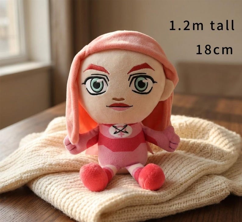 May include: A plush doll with a pink and red outfit, featuring a stylized face with large green eyes and pink hair. The doll is approximately 18 cm tall and 1.2 meters tall. The doll is sitting on a cream-colored knitted blanket.
