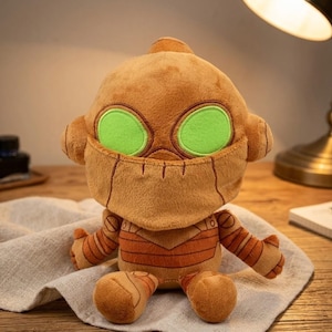 May include: A plush toy robot with a brown body, green eyes, and stitched details. The robot has a rounded head, small ears, and striped arms and legs. It is sitting on a light-colored cloth.