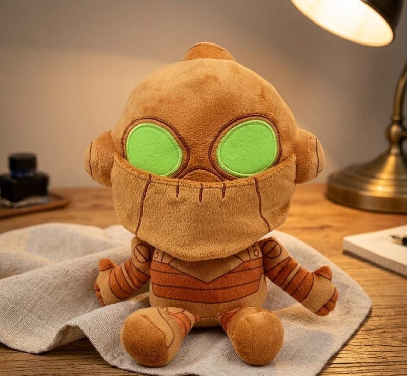 May include: A plush toy robot with a brown body, green eyes, and stitched details. The robot has a rounded head, small ears, and striped arms and legs. It is sitting on a light-colored cloth.