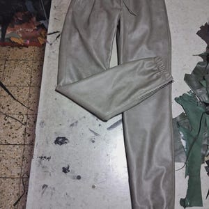 May include: Gray leather pants with a drawstring waist and elastic cuffs. The pants have pockets and a zipper detail at the ankle. The leather appears soft and has a matte finish. The pants are laid out on a light-colored surface.