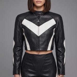 May include: A cropped black leather jacket featuring a prominent white chevron pattern across the chest and sleeves. The jacket has a high collar with a snap button closure, a central zipper, and long sleeves. A silver triangular pendant necklace is worn.