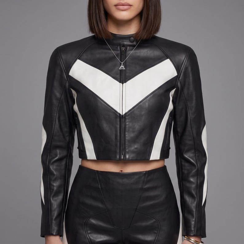 May include: A cropped black leather jacket featuring a prominent white chevron pattern across the chest and sleeves. The jacket has a high collar with a snap button closure, a central zipper, and long sleeves. A silver triangular pendant necklace is worn.