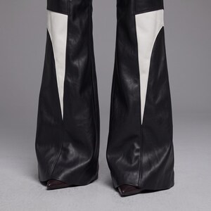May include: Black faux leather flared trousers featuring large, inverted white triangular panels on the outer legs. The pants have a smooth, matte finish and a wide-leg silhouette that extends down to cover dark, pointed-toe footwear at the base.