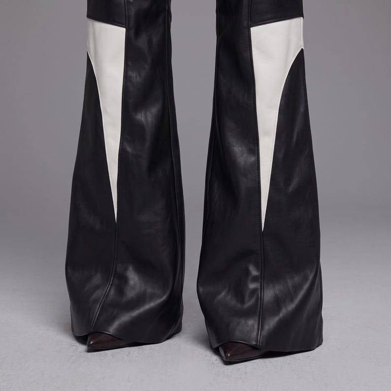 May include: Black faux leather flared trousers featuring large, inverted white triangular panels on the outer legs. The pants have a smooth, matte finish and a wide-leg silhouette that extends down to cover dark, pointed-toe footwear at the base.