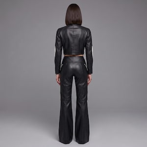 May include: A black leather outfit consisting of a cropped long-sleeve jacket and high-waisted flared trousers. The jacket features white piping along the sleeves, while the trousers include white piping details along the hips and legs.