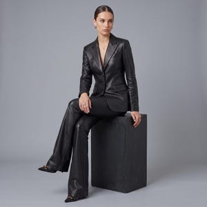 K&ouml;nnte beinhalten: Ein schwarzer Lederblazer mit steigendem Revers, zwei Kn&ouml;pfen auf der Vorderseite und zwei Pattentaschen, kombiniert mit einer passenden, ausgestellten Lederhose in Schwarz. Das Set besticht durch eine glatte Oberfl&auml;che und einen strukturierten Schnitt.