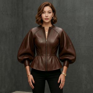 Könnte beinhalten: Eine dunkelbraune Lederjacke mit durchgehendem Frontreißverschluss, tailliertem Schnitt und voluminösen Puffärmeln. Die Jacke hat ein strukturiertes Oberteil mit sichtbaren Nahtdetails und wird mit einer schwarzen Hose kombiniert.