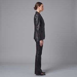 K&ouml;nnte beinhalten: Ein schwarzer Lederblazer mit steigendem Revers und drei Kn&ouml;pfen auf der Vorderseite, kombiniert mit einer passenden schwarzen Lederhose. Das Set hat eine glatte, polierte Oberfl&auml;che, lange &Auml;rmel und eine taillierte Passform, Seitenansicht.