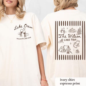 May include: Ivory-colored t-shirt with espresso-colored print. The front features "Lake Crew" text and wine glasses. The back displays "The Wilson Lake Trip" with camping and lake-themed illustrations. The shirt is a casual, unisex style.