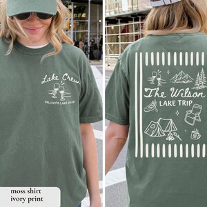 May include: A moss green t-shirt with white text and graphics. The front of the shirt says "Lake Crew" with wine glasses and "Vallecito Lake 2026". The back says "The Wilson Lake Trip" with camping-themed illustrations.