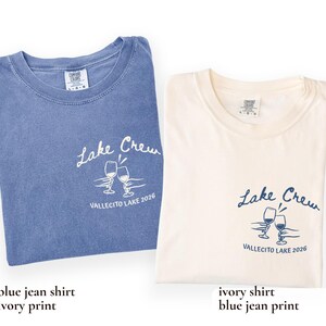 May include: Two folded t-shirts, one blue and one ivory, each with the text "Lake Crew" and "VALLECITO LAKE 2026" printed on them. The design includes two wine glasses. The blue shirt has an ivory print, and the ivory shirt has a blue print.