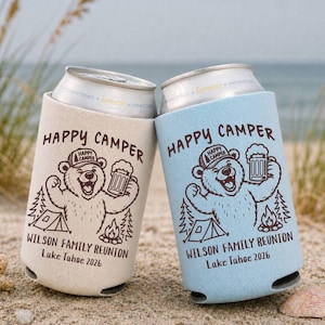 May include: Two foam can coolers, one beige and one light blue, each featuring a brown graphic of a bear wearing a hat, holding a beer mug, next to a tent and campfire. The text reads Happy Camper, Wilson Family Reunion, and Lake Tahoe 2026.