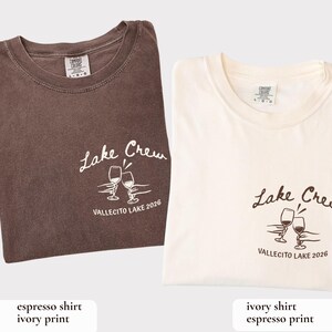 May include: Two folded t-shirts, one espresso-colored and one ivory, each with a crew neck. Both shirts feature the text "Lake Crew" and a graphic of two wine glasses, with "VALLECITO LAKE 2026" below.