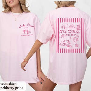 May include: Light pink t-shirt with the text "Lake Crew" and "Vallecito Lake 2020" in pink script on the front. The back of the shirt features a pink graphic with the text "The Wilson Lake Trip" and camping-themed illustrations.