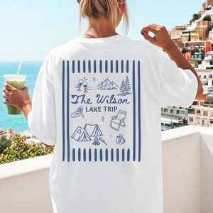 May include: White t-shirt with a blue graphic on the back. The graphic includes the text "The Wilson Lake Trip" with illustrations of mountains, trees, a tent, a canoe, and other camping-themed images. The shirt is a relaxed fit.