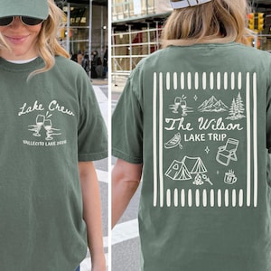 Puede incluir: Camiseta verde salvia con texto gráfico e ilustraciones en blanco. La parte delantera de la camiseta dice "Lake Crew Vallecito Lake 2026" con copas de vino. La parte trasera presenta un diseño temático de camping con el texto "The Wilson Lake Trip".