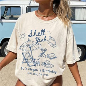 Puede incluir: Camiseta color crema con un gráfico azul con temática de playa. El diseño incluye un sol, una concha, una sombrilla, una silla, un sombrero y el texto "Shell Yeah". También está presente el texto "It's Megan's Birthday, Malibu, CA 2026".