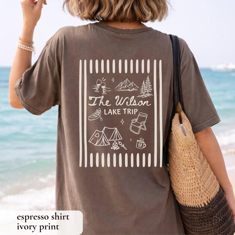 May include: Espresso-colored t-shirt with an ivory print featuring the text "The Wilson Lake Trip" and camping-themed illustrations. The design includes a tent, mountains, and a canoe. The shirt is a relaxed fit.