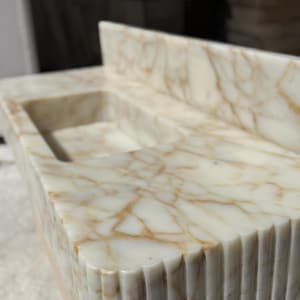 May include: A rectangular marble sink with a built-in backsplash. The sink basin is recessed, and the marble features a cream and gold veining pattern. The front edge has a fluted design, adding texture to the stone.