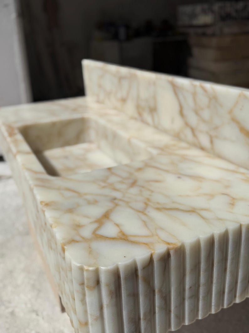 May include: A rectangular marble sink with a built-in backsplash. The sink basin is recessed, and the marble features a cream and gold veining pattern. The front edge has a fluted design, adding texture to the stone.