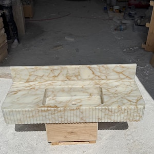 May include: A rectangular marble vanity with a built-in sink. The vanity has a cream-colored base with gold veining and a fluted front. The sink is rectangular and centered. The vanity is supported by a wooden base.