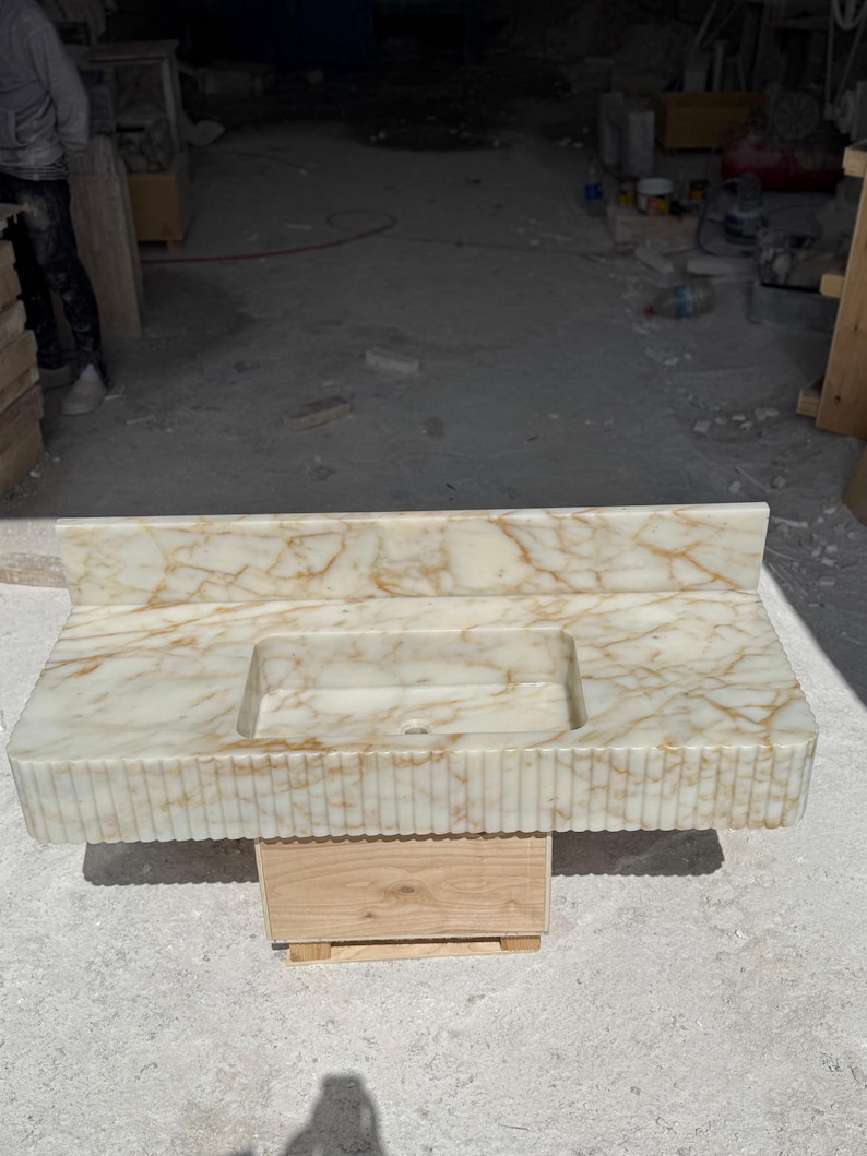 May include: A rectangular marble vanity with a built-in sink. The vanity has a cream-colored base with gold veining and a fluted front. The sink is rectangular and centered. The vanity is supported by a wooden base.