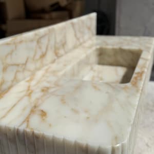 May include: A close-up of a marble countertop with a built-in sink. The countertop is white with brown veining and a textured, grooved edge. The sink is rectangular and has a smooth interior.