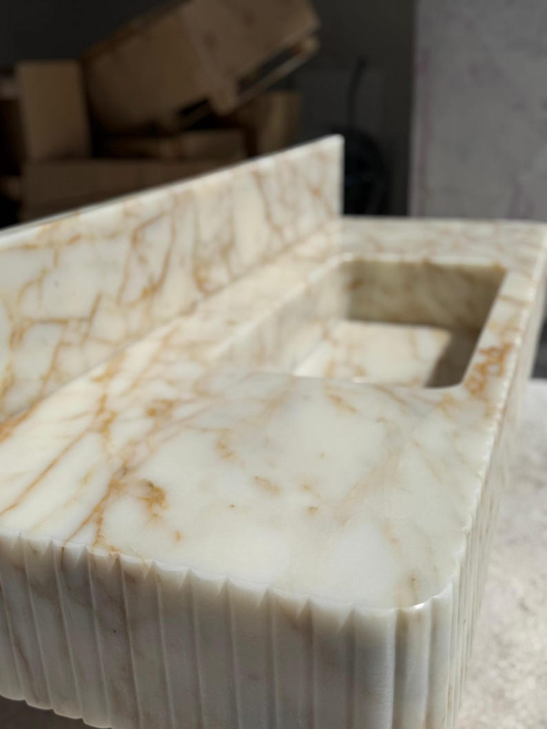 May include: A close-up of a marble countertop with a built-in sink. The countertop is white with brown veining and a textured, grooved edge. The sink is rectangular and has a smooth interior.