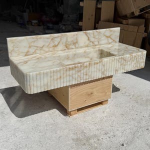 May include: A marble vanity with a rectangular sink basin and a backsplash. The vanity has a light beige color with brown veining and a fluted front. It is mounted on a wooden base.