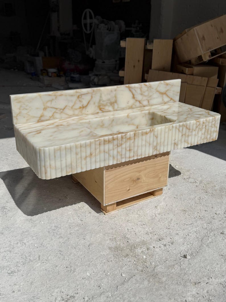 May include: A marble vanity with a rectangular sink basin and a backsplash. The vanity has a light beige color with brown veining and a fluted front. It is mounted on a wooden base.