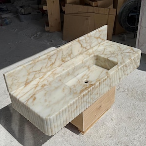 May include: A rectangular marble sink with a built-in basin and a backsplash. The sink has a cream-colored base with gold veining and a fluted edge. It is mounted on a light-colored wooden base.