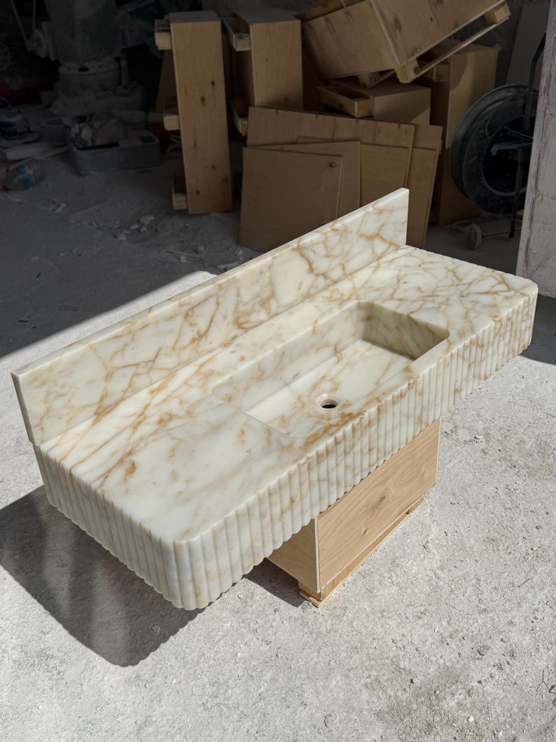 May include: A rectangular marble sink with a built-in basin and a backsplash. The sink has a cream-colored base with gold veining and a fluted edge. It is mounted on a light-colored wooden base.