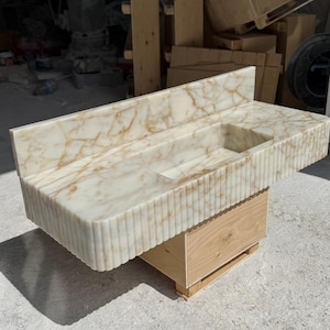 May include: A marble vanity with a rectangular sink basin. The countertop and backsplash feature a cream and gold veined pattern. The front of the vanity has a vertical ribbed design. It rests on a wooden base.