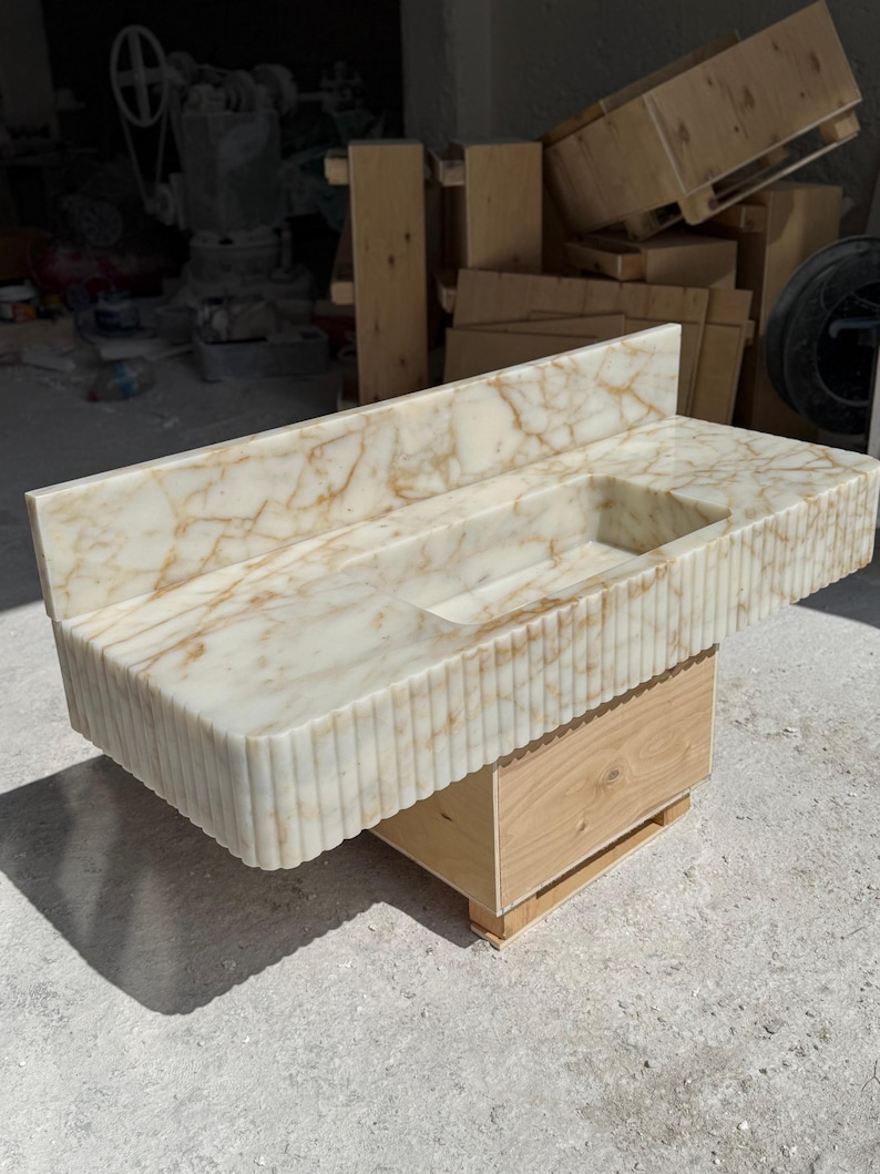 May include: A marble vanity with a rectangular sink basin. The countertop and backsplash feature a cream and gold veined pattern. The front of the vanity has a vertical ribbed design. It rests on a wooden base.