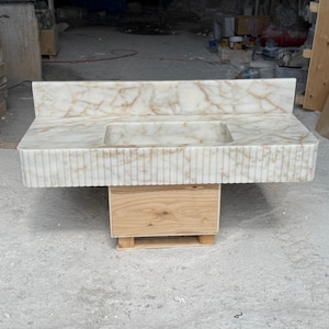 May include: A white marble vanity with a rectangular integrated sink. The countertop has brown veining and a backsplash. The front of the vanity has vertical grooves. It is supported by a wooden cabinet on a small pallet.