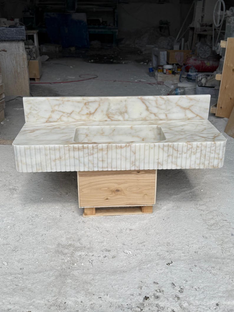 May include: A white marble vanity with a rectangular integrated sink. The countertop has brown veining and a backsplash. The front of the vanity has vertical grooves. It is supported by a wooden cabinet on a small pallet.