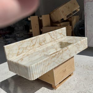 May include: A marble bathroom sink with a rectangular basin and a backsplash. The sink is off-white with brown veining and a vertically ribbed front. It sits on a light wood base.