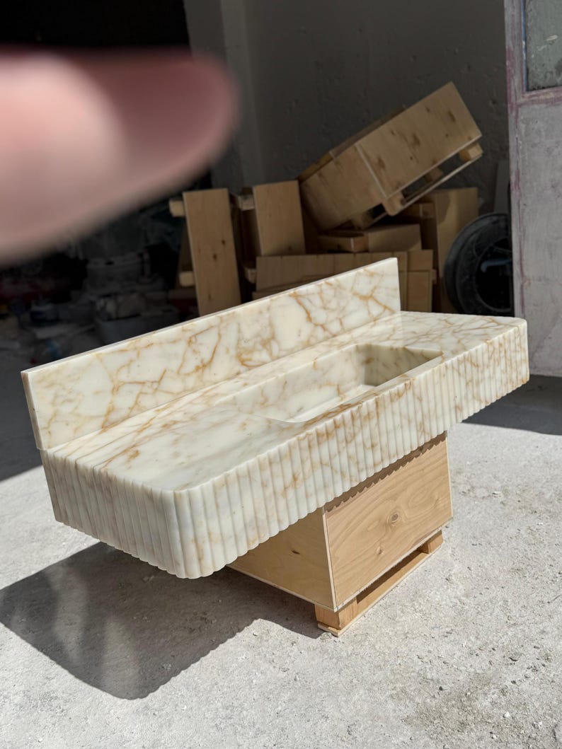 May include: A marble bathroom sink with a rectangular basin and a backsplash. The sink is off-white with brown veining and a vertically ribbed front. It sits on a light wood base.