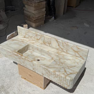 May include: A rectangular, cream-colored marble sink with a built-in basin and backsplash. The sink has a veined pattern in shades of gold and brown. The front edge features vertical grooves. It is supported by a wooden base.