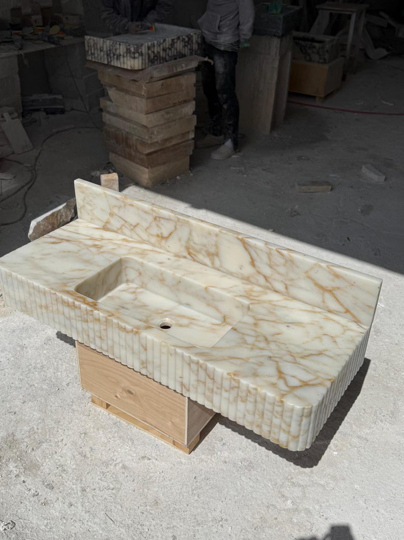 May include: A rectangular, cream-colored marble sink with a built-in basin and backsplash. The sink has a veined pattern in shades of gold and brown. The front edge features vertical grooves. It is supported by a wooden base.
