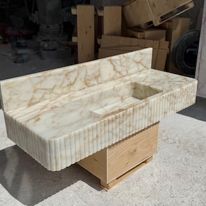 May include: A rectangular marble sink with a built-in backsplash and a carved, fluted front. The sink has a light beige color with brown veining and sits atop a wooden base. The sink's dimensions are approximately 48 inches long, 20 inches wide, and 10 inches high.