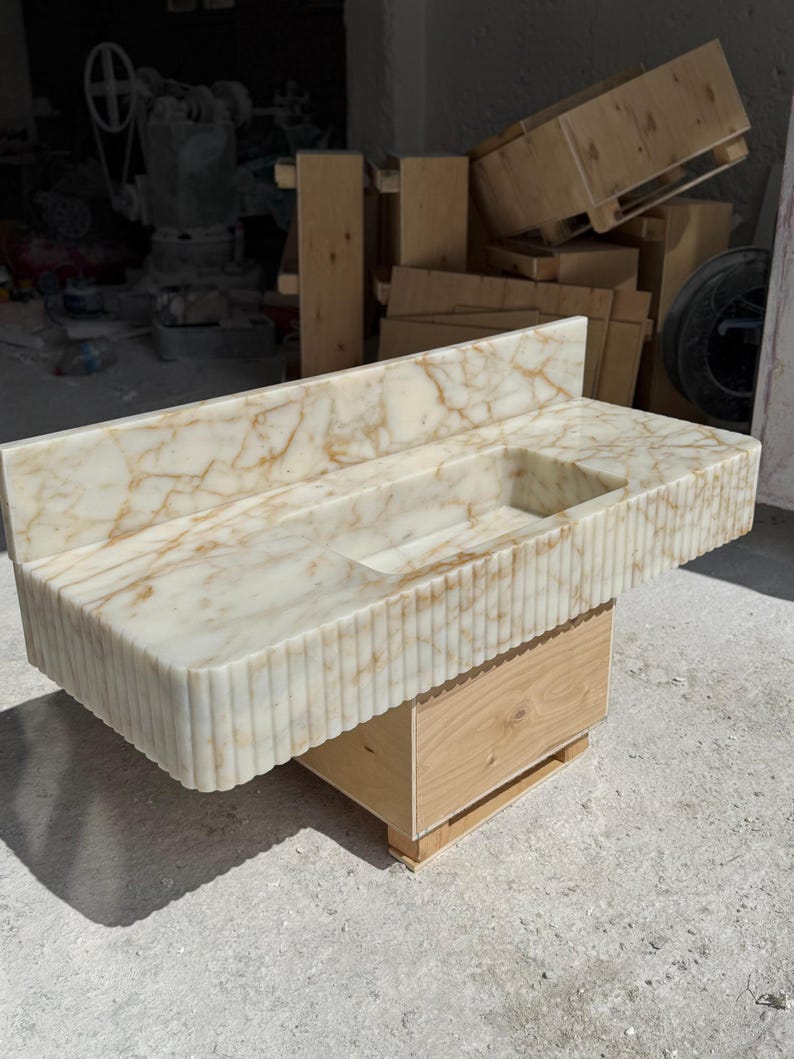 May include: A rectangular marble sink with a built-in backsplash and a carved, fluted front. The sink has a light beige color with brown veining and sits atop a wooden base. The sink's dimensions are approximately 48 inches long, 20 inches wide, and 10 inches high.