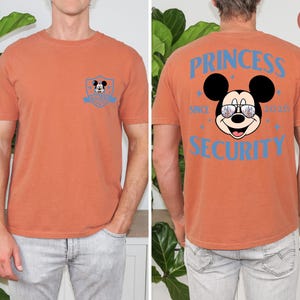 May include: Burnt orange t-shirt with a Mickey Mouse graphic on the front and back. The front features a small Mickey Mouse emblem. The back displays Mickey wearing sunglasses with the text "PRINCESS SECURITY SINCE 2020".