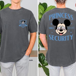 May include: A charcoal gray t-shirt featuring a Mickey Mouse design. The front has a small emblem with Mickey. The back displays Mickey wearing sunglasses, with the words "PRINCESS SECURITY SINCE 2026" in blue.