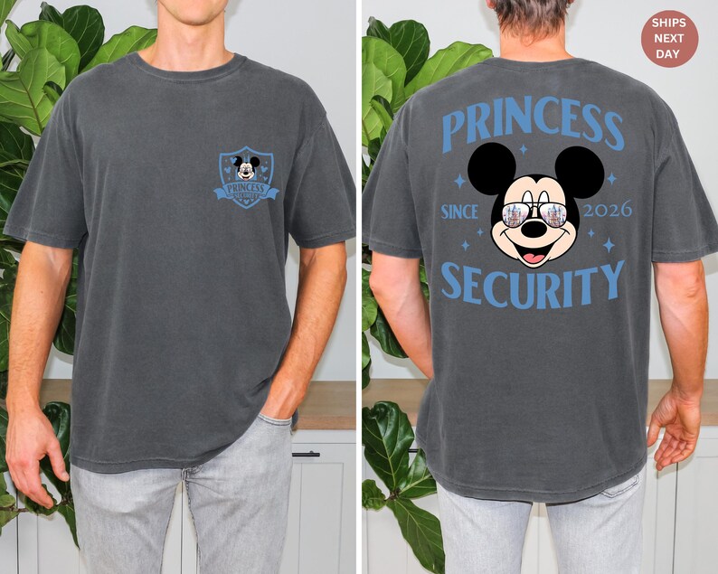 May include: A charcoal gray t-shirt featuring a Mickey Mouse design. The front has a small emblem with Mickey. The back displays Mickey wearing sunglasses, with the words "PRINCESS SECURITY SINCE 2026" in blue.