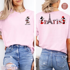 May include: Light pink t-shirt featuring a Minnie Mouse graphic with the text "YOUR NAME" on the front. The back of the shirt displays Mickey and Minnie Mouse with the Eiffel Tower and the word "Paris".