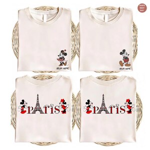 May include: Four cream-colored t-shirts with cartoon character designs. Two shirts feature Mickey and Minnie Mouse, and the other two have the Eiffel Tower and the word "Paris". Each shirt has the text "YOUR NAME".
