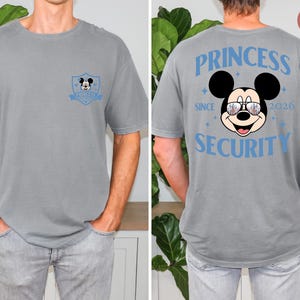 May include: Gray t-shirt featuring a Mickey Mouse graphic on the front and back. The front has a small emblem with Mickey. The back displays "PRINCESS SECURITY SINCE 2026" with Mickey wearing sunglasses. A fun, casual tee.